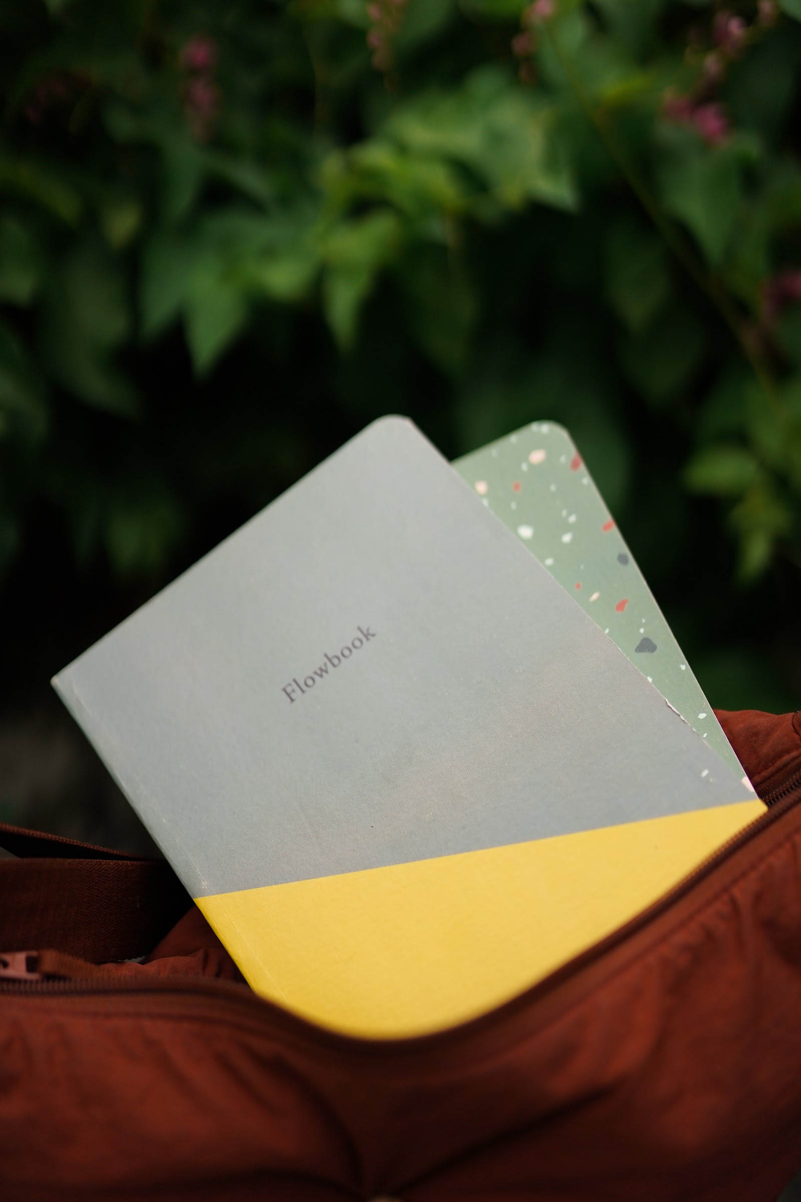 Two notebooks, one grey and yellow, one green terrazzo, tucked into a rust-red bag outdoors