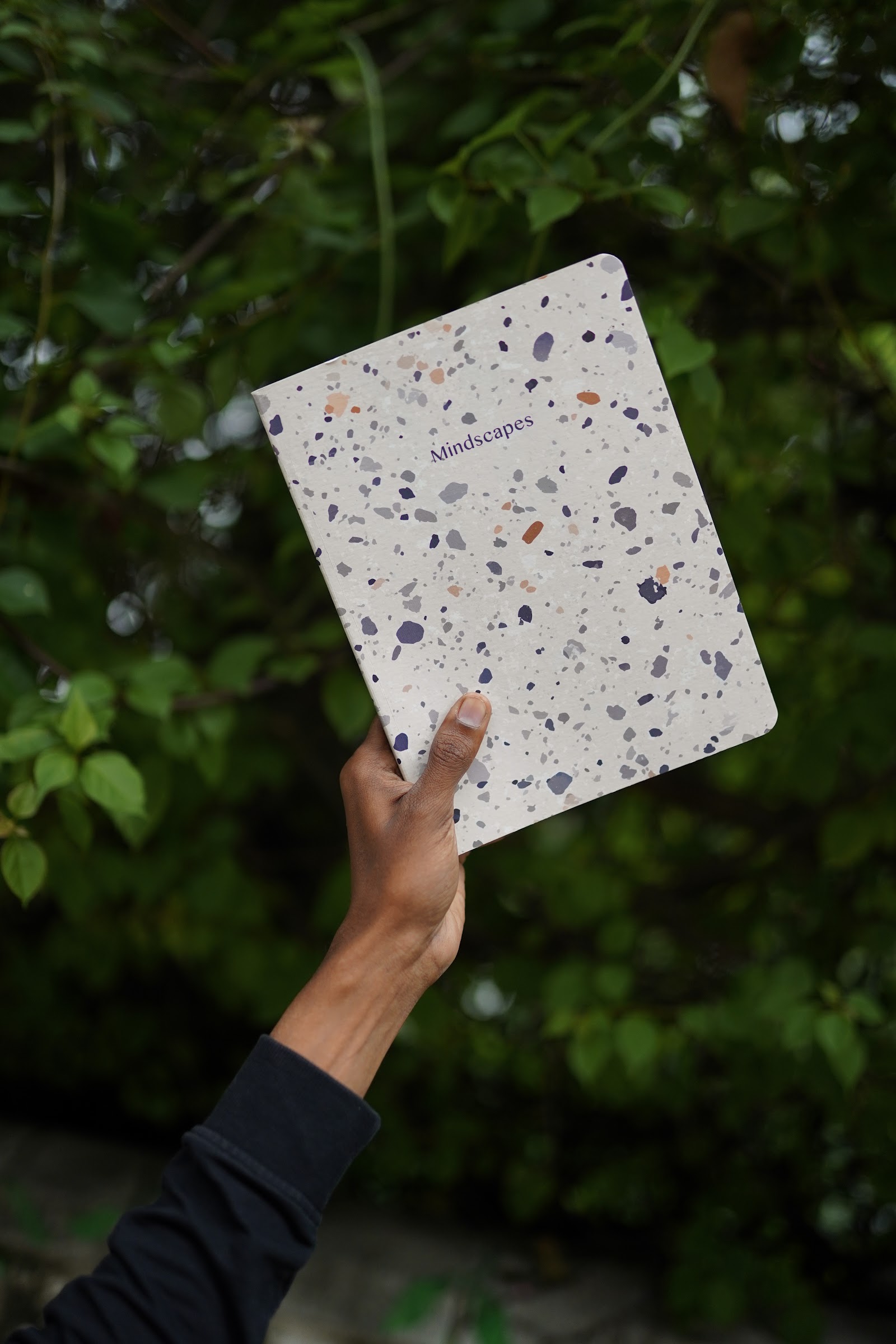 A hand holding up the Mindscapes terrazzo-print notebook against green foliage